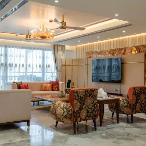 A backlit onyx slab in a brass frame wraps the TV panel of a large living room, with two dark floral wing chairs facing blush sofas