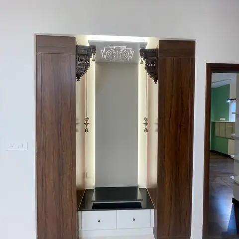 A backlit pooja alcove framed by dark wood pillars with carved elephant brackets and hanging brass bell-lamps, above a black-stone platform and white drawers.