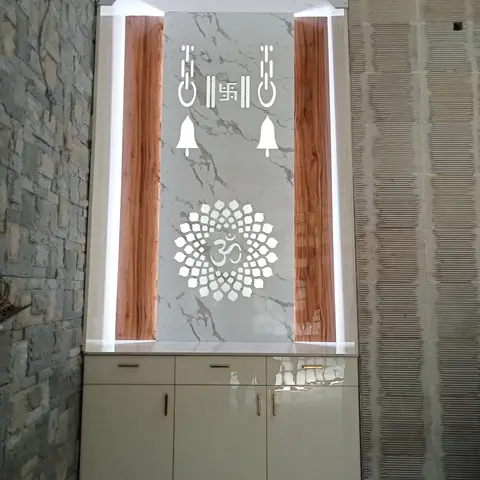 A backlit pooja unit with a marble panel featuring bells, a swastik, and an Om motif flanked by wood pillars, with glossy storage cabinets below.