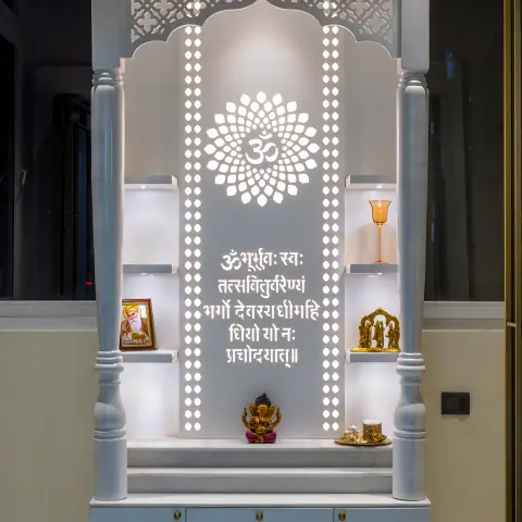 A backlit pooja unit with a carved jaali arch, a Gayatri Mantra text panel, brass idol niches on either side and a marble base cabinet with brass knob hardware.