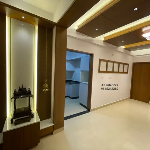 A backlit wooden pooja niche with hanging brass bells sits beside an open hallway to the kitchen, under a wood-panelled cove-lit ceiling.