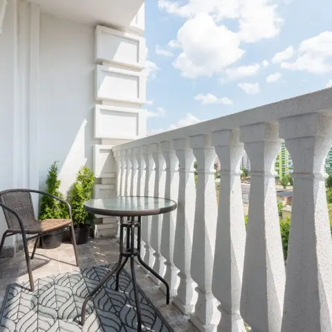 A balcony with a wicker chair and round glass bistro table on a patterned rug, framed by a decorative stone balustrade overlooking the city skyline.