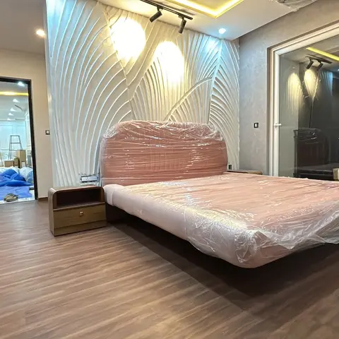 A bedroom mid-handover with a sculptural white fluted 3D wall panel behind a floating platform bed still wrapped in protective plastic
