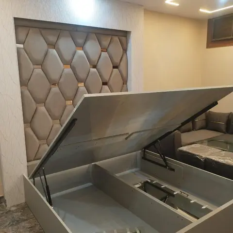 A bedroom mid-setup with a grey hydraulic storage bed lifted open, in front of a diamond-tufted upholstered headboard wall niche