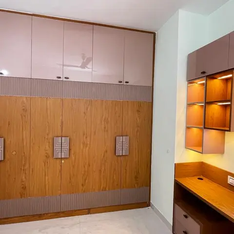 A bedroom's study corner with a built-in wood desk under a backlit display cubby, beside a large two-tone mauve-and-wood wardrobe wall.