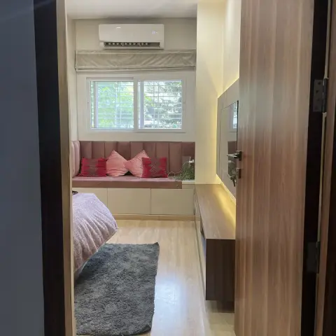 A bedroom viewed through the doorway, with a mauve channel-tufted window seat bench topped with pink and red cushions, a grey shag rug