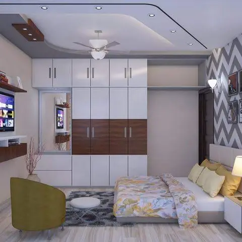 A bedroom where a white-and-walnut wardrobe wall with a mirrored dresser niche faces a grey chevron wallpaper wall hung with black-framed prints