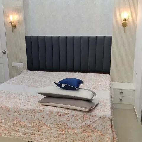 A bedroom with a built-in channel-tufted navy headboard wall flanked by fluted pilasters and brass wall sconces, floral wallpaper