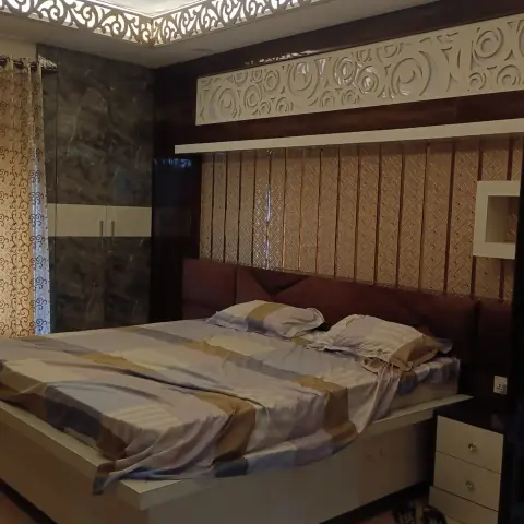 A bedroom with a carved scrollwork jaali trim framing a cove-lit ceiling, a tufted maroon headboard against a fluted gold panel wall