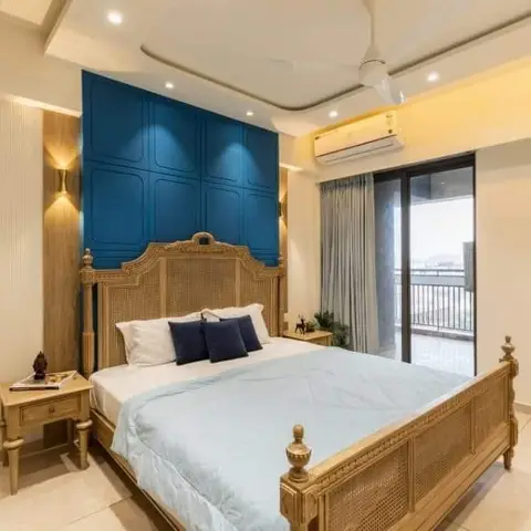A bedroom with a carved wood cane bed against a deep teal panelled feature wall, cove-lit ceiling, and a glass sliding door opening to a private balcony.