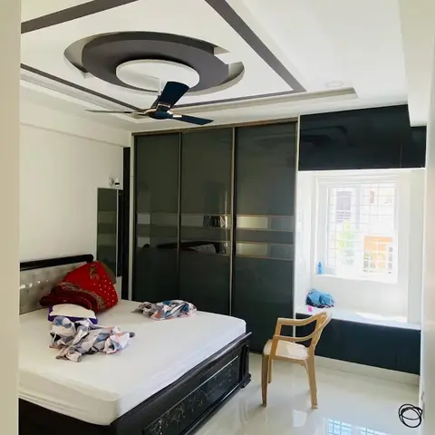 A bedroom with a circular POP false-ceiling ring around the fan, a black glossy sliding wardrobe with mirror-strip inlays, and a carved dark-wood bed frame
