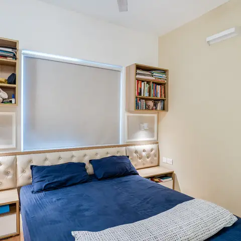 A bedroom with a cream diamond-tufted upholstered headboard flanked by wall-mounted bookshelves and sconce reading lights, and a navy bed cover.