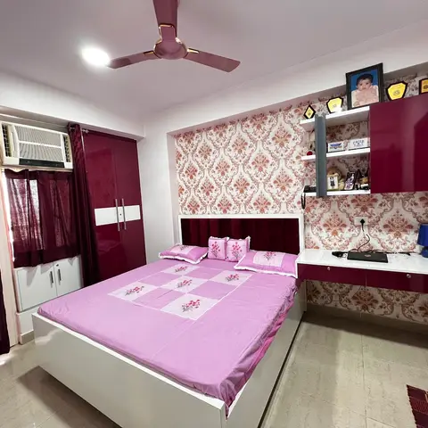 A bedroom with a damask-patterned maroon wallpaper headboard wall, glossy maroon wardrobe, pink floral bedding, and a built-in study desk with open shelving.