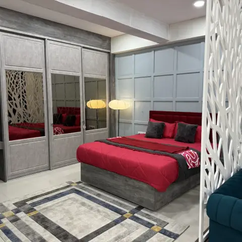 A bedroom with a deep red channel-tufted headboard and quilted bedcover, a mirrored sliding wardrobe, a decorative white jaali room divider