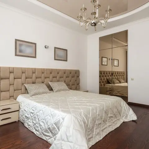 A bedroom with a full-wall tufted beige upholstered headboard, a crystal chandelier hung from a glossy curved cove ceiling, a mirrored wardrobe