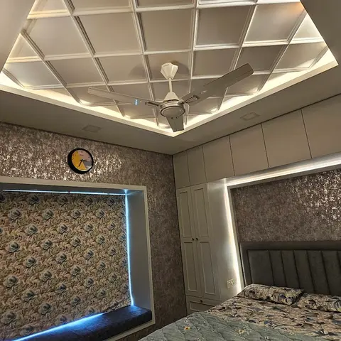 A bedroom with a geometric coffered cove-lit false ceiling over a ceiling fan, metallic textured wallpaper, a floral-upholstered window seat nook