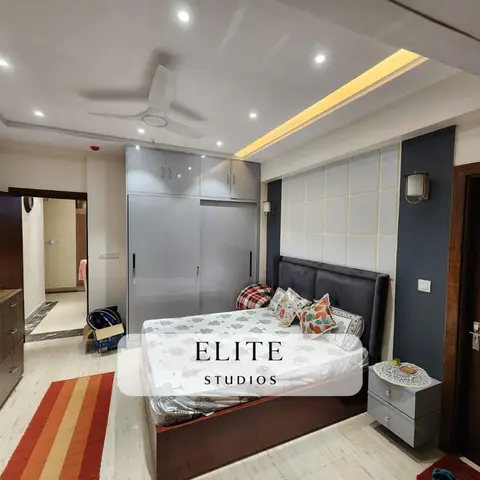A bedroom with a glossy grey sliding-door wardrobe and a slate-blue tiled panel headboard wall lit by cove ceiling lighting and brass wall sconces.