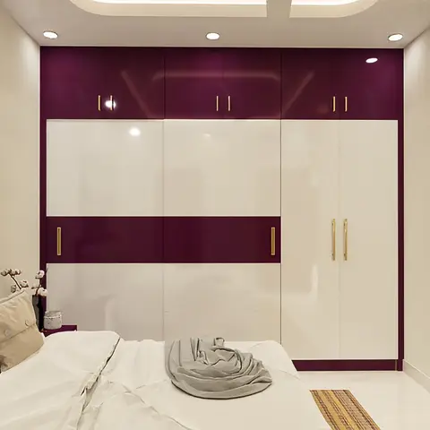 A bedroom with a glossy maroon-and-white wardrobe with gold handles, a tufted headboard bed, and roman blinds at the window.