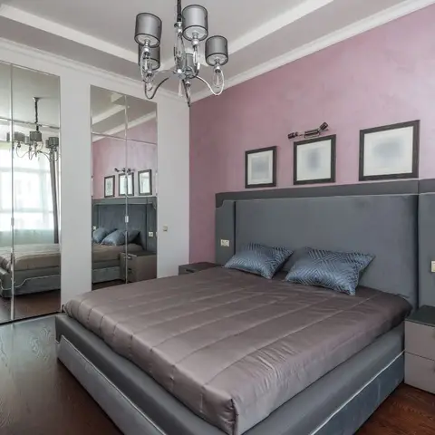 A bedroom with a grey velvet channel-tufted headboard against a rose-pink textured accent wall, a mirrored wardrobe reflecting the room