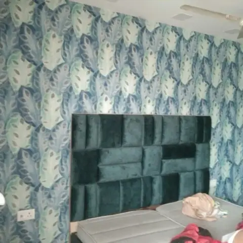 A bedroom with a leaf-print teal wallpaper wall behind a channel-tufted emerald velvet headboard, photographed with clothes scattered on the unmade bed.
