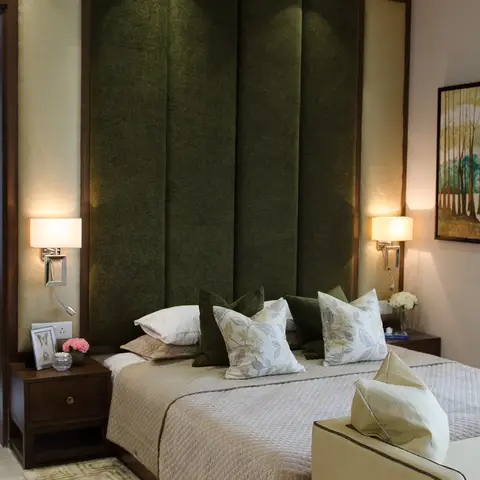 A bedroom with a tall channel-tufted olive velvet headboard framed in dark wood, brass reading sconces on either side