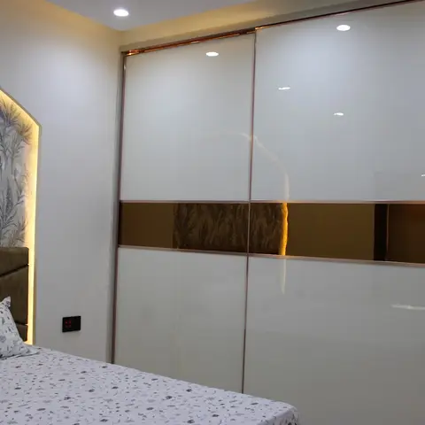 A bedroom with a tufted brown headboard set in a cove-lit arched wallpaper niche beside a glossy white sliding wardrobe with a bronze mirror strip