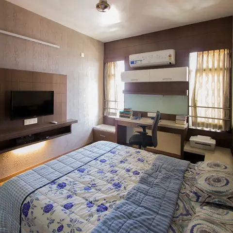 A bedroom with a wall-mounted TV on a wood-panel unit, a built-in study desk with overhead cabinets and AC, and a leather headboard bed with a floral quilt.