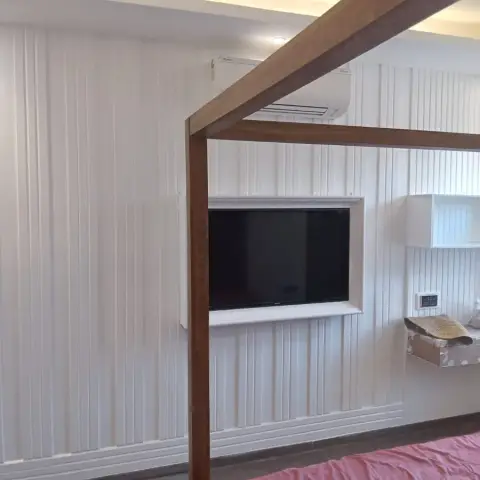A bedroom with a white fluted-panel accent wall, a wood-framed TV niche recessed into the panelling, a wall-mounted split AC, cove ceiling lighting