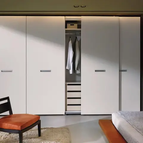 A bedroom with a white hinged wardrobe wall — one door open to reveal a hanging rail and drawer stack — a mid-century-style chair with a burnt-orange leather