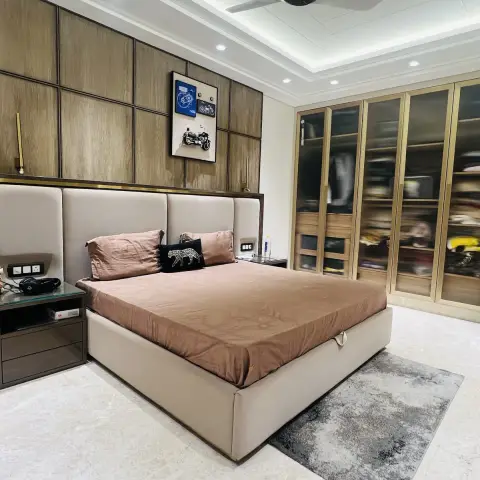 A bedroom with a wood-grid panelled and brass-trimmed headboard wall, a glass-front wardrobe, a framed motorcycle print, and a grey rug on marble flooring.