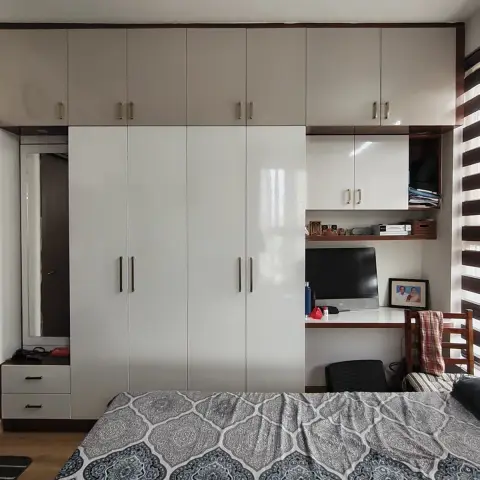 A bedroom with floor-to-ceiling glossy white wardrobes with an overhead loft, a built-in study desk with computer monitor