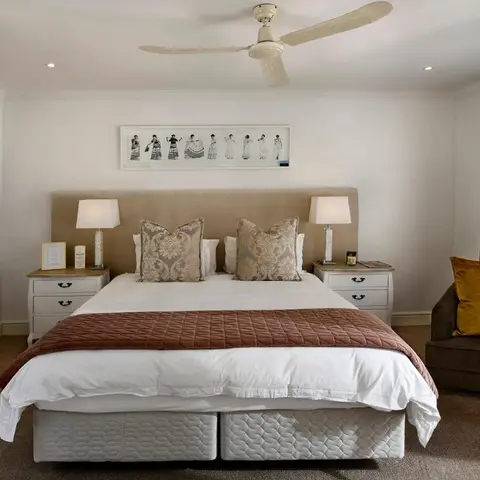 A beige upholstered-headboard bed flanked by matching nightstands and lamps, with a brown armchair and mustard cushion in the corner.