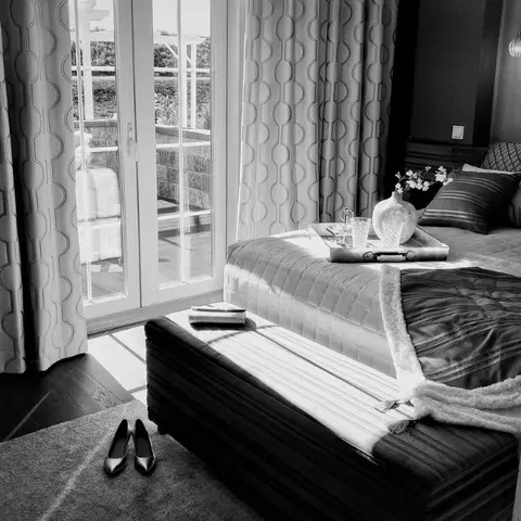 A black-and-white shot of a styled bedroom: breakfast tray and vase on a quilted bed, a ribbed ottoman bench across its foot