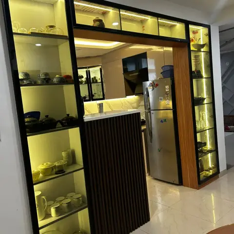 A black-framed backlit glass crockery display flanks a fluted wood-panelled doorway opening into a marble-backsplash kitchen with a steel refrigerator.
