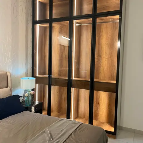 A black steel-framed glass wardrobe with LED-lit oak-veneer interior along a botanical-print wallpapered wall in a bedroom.