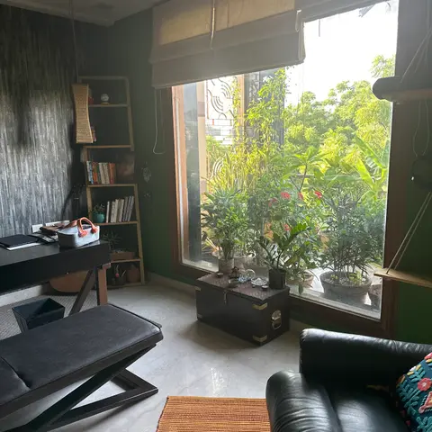 A boho home study with a dark textured accent wall, black leather sofa and bench, a wooden bookshelf, and a large window framed by potted plants.