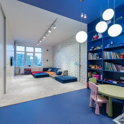 A bold blue kids' playroom with a full-height built-in bookshelf packed with toys and books, low colorful seating and globe pendant lights.
