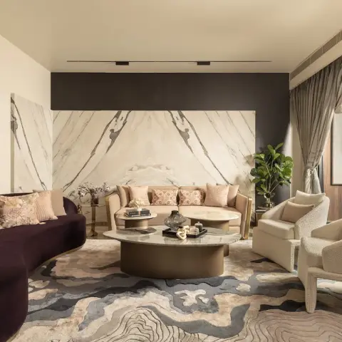 A book-matched cream marble slab wall rises behind a blush gold-framed settee, faced across bronze-drum marble tables by a deep aubergine curved velvet sofa