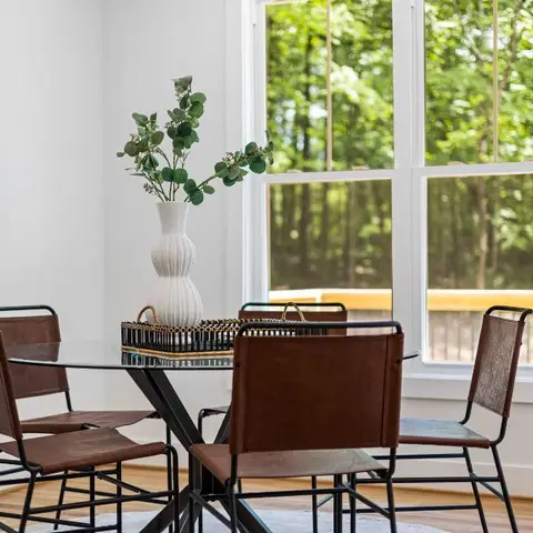 A bright dining nook with a round glass-top table on a black metal base, brown leather-and-metal chairs, and a ribbed white vase of eucalyptus