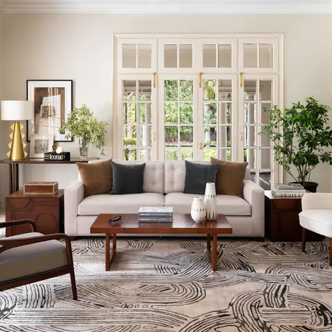 A bright living room with a cream tufted sofa, a wood-and-cane mid-century armchair, a bold black-and-white swirl-pattern area rug