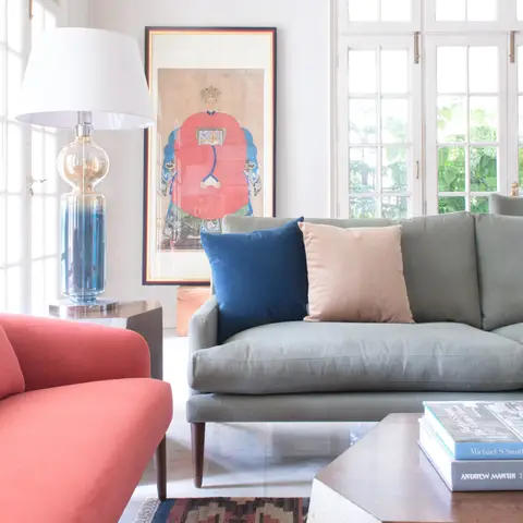 A bright living room with a coral velvet sofa facing a sage-green sofa, styled with mixed jewel-tone cushions beneath a framed portrait artwork.