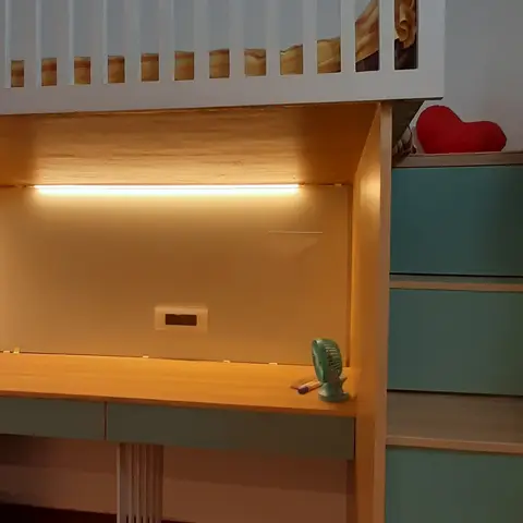 A built-in loft bed with a slatted white guardrail sits above a wood-toned study desk lit by an under-shelf LED strip, paired with mint-green storage drawers.
