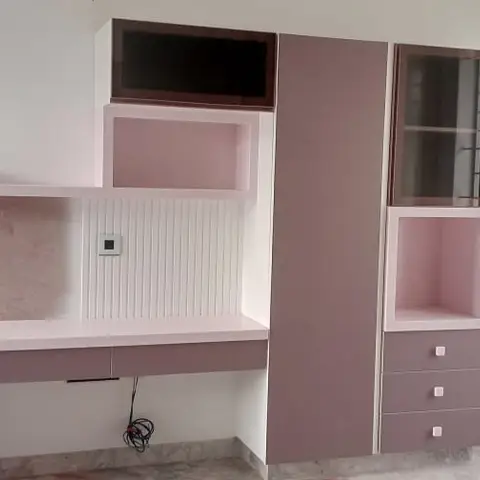 A built-in study desk and wardrobe combination in soft mauve and white, with open display shelving and a fluted white backsplash beside a window with iron
