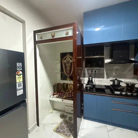 A built-in wood-framed pooja alcove with deity photos and brass lamps set beside a glossy blue modular kitchen with a stone-topped gas hob.