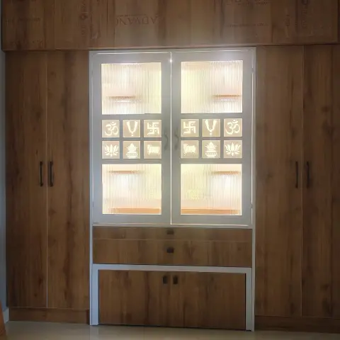A built-in wooden pooja unit with backlit fluted-glass doors featuring Om, swastik and lotus motifs, flanked by full-height wardrobe cabinets