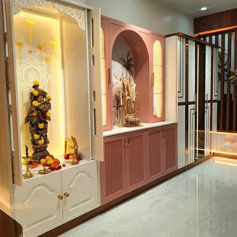 A carved white pooja cabinet with backlit Om motif stands beside a dusty-pink fluted-arch display unit in a home entrance passage.