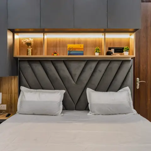 A chevron channel-tufted grey headboard bed with a backlit wood display niche and overhead storage cabinets, flanked by a matching wood-finish door.