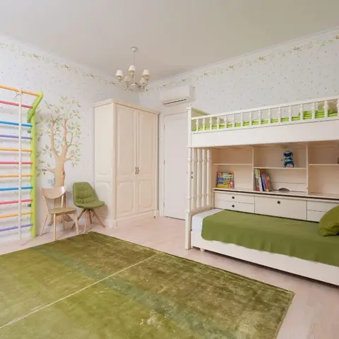 A child's bedroom with a white built-in bunk bed dressed in green bedding, a tree-mural accent wall, and a colourful indoor climbing wall.