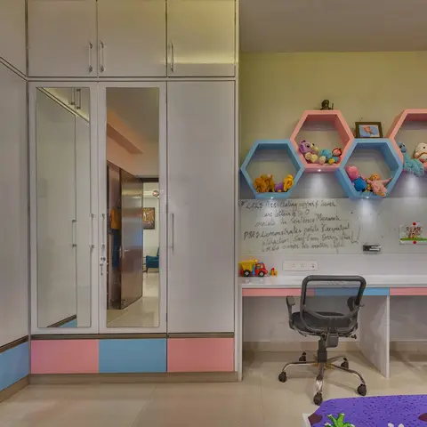 A child's bedroom with a pink-and-blue two-tone wardrobe, hexagonal display shelves holding stuffed toys, and a study desk against a whiteboard wall.