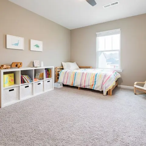 A child's bedroom with a striped rainbow-colored bedspread, cubby storage bins filled with books and toys, dinosaur wall art and a wooden dollhouse.